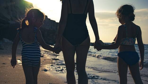 Мама и две дочки гуляют по берегу моря в купальниках, держась за руки