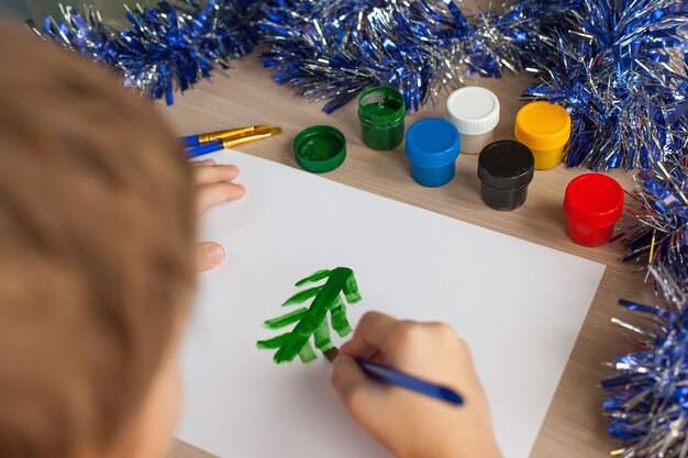 Мальчик рисует елку и снег на листе бумаги детский новогодний рисунок гуашь и кисточки развитие ребенка