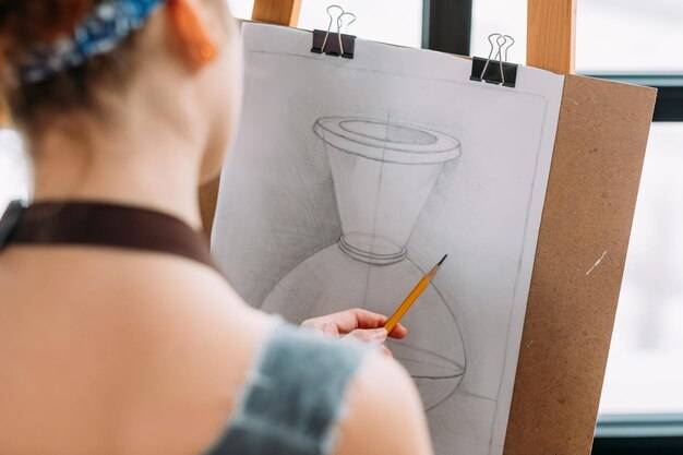 Художественная студия крупным планом вид сзади художника-левши, измеряющего пропорции карандашом в вазе для рисования