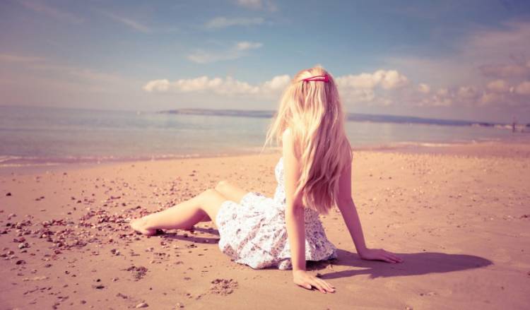 Фото со спины блондинки в платье, которая сидит на берегу моря